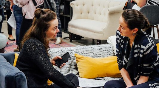 Two women chat together seated on a couch in the middle of a cocktail party.