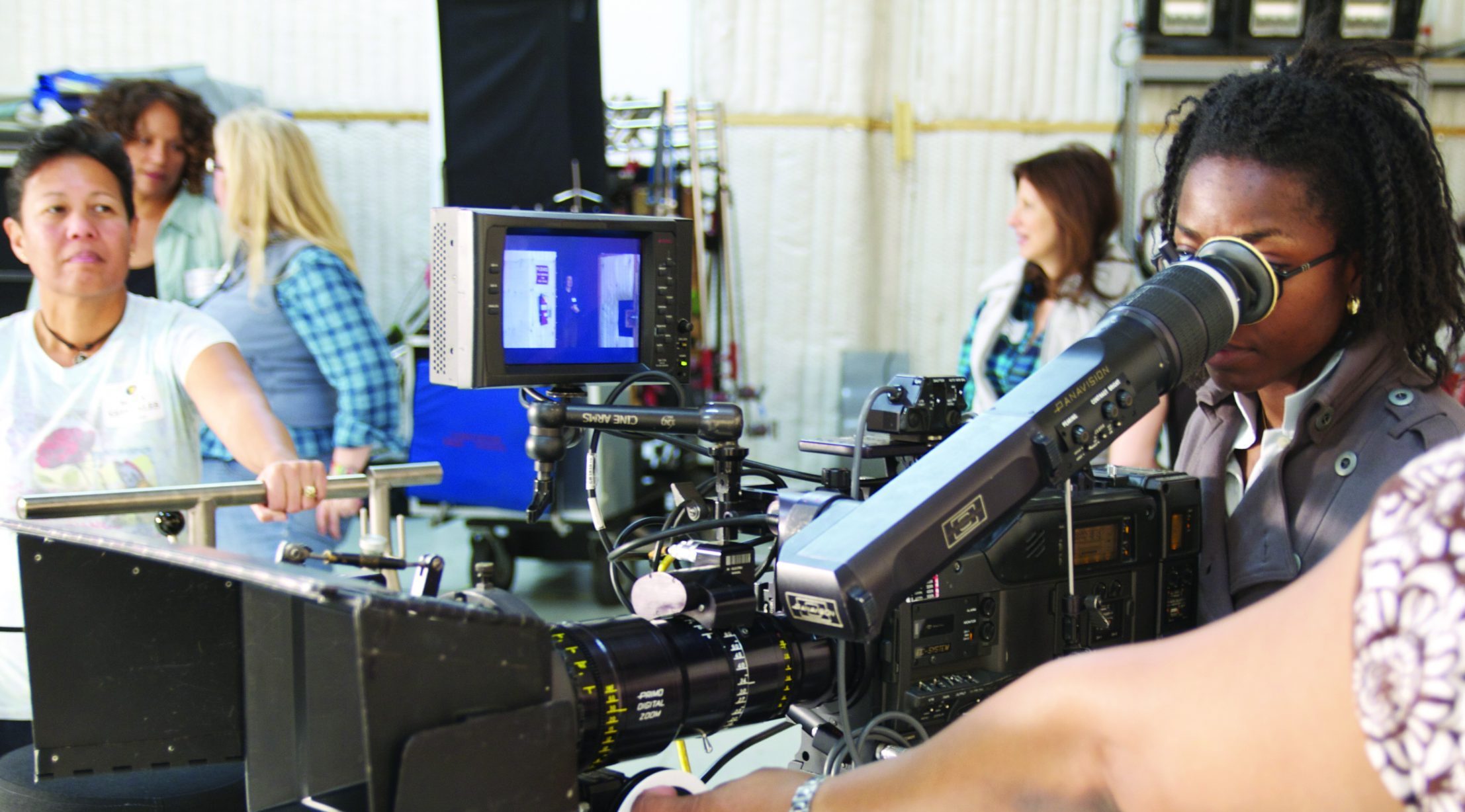 A woman peers into a camera eye piece, with other women crew members in the background.