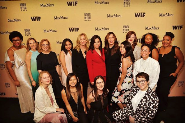 Sixteen sharply dressed women pose in front of a photo backdrop on a red carpet.